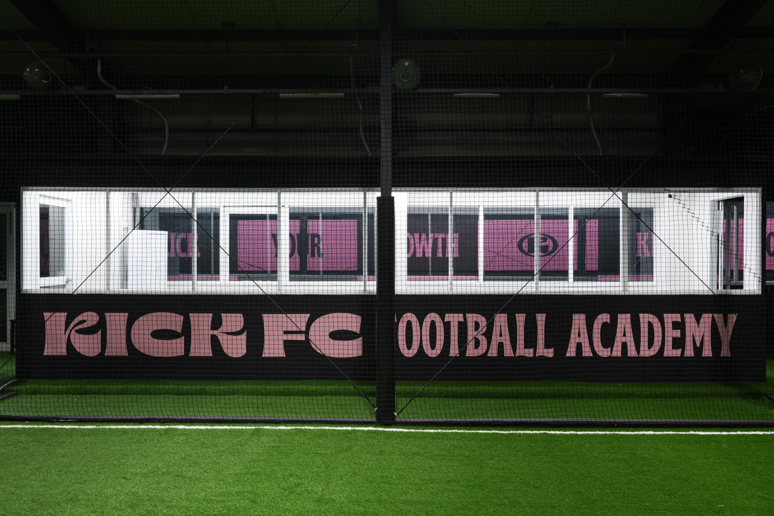그물 너머로 보이는 벽면 사이니지. 분홍색 창문에 KICK YOUR GROWTH 문구가, 아래 검은 벽면에 KICK FC FOOTBALL ACADEMY 글자가 적혀 있다.